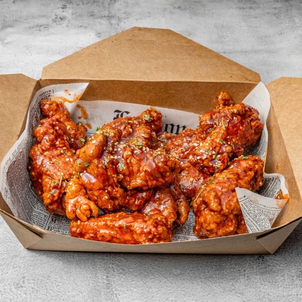 DAK Korean Fried Chicken wings, coated in red sauce and topped with herbs, in a takeout box.
